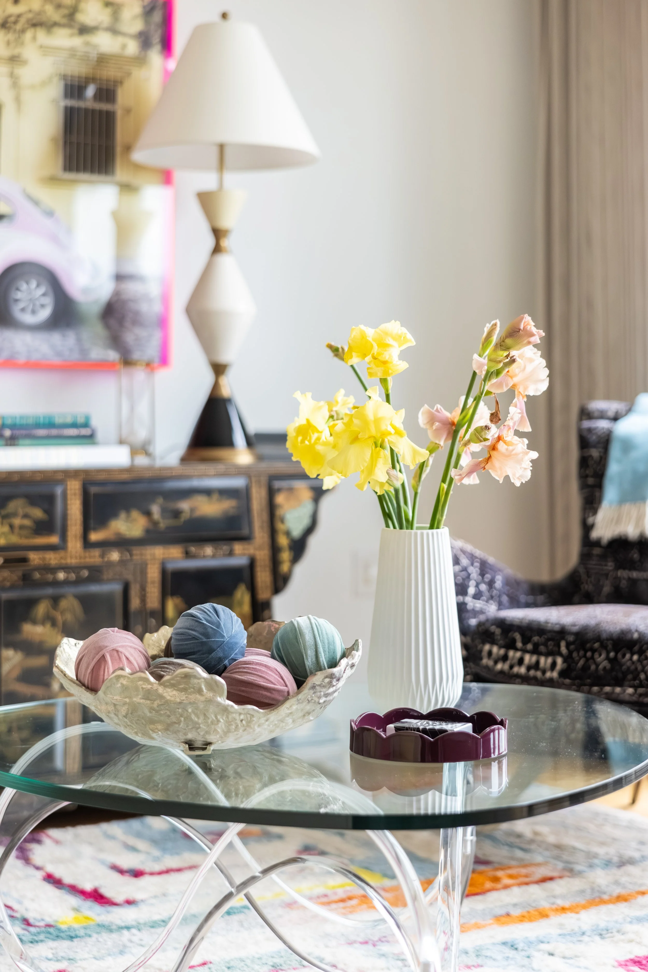 A clear glass table with a decorative ceramic bowl filled with pastel-colored yarn balls, a purple and white ceramic container, and a white vase with yellow and pink flowers. In the background, there is a lamp, a Chinese-style cabinet, a couch, and a framed picture on the wall.