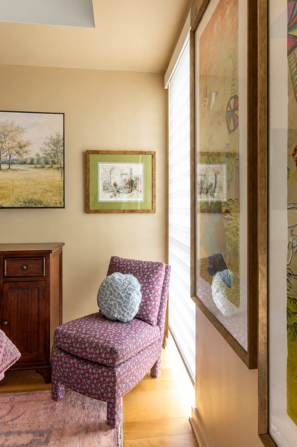 Interior corner of a cozy room with a pink patterned chair with a round floral pillow, a wooden cabinet, framed artwork on a beige wall, and a large window with architectural blinds.