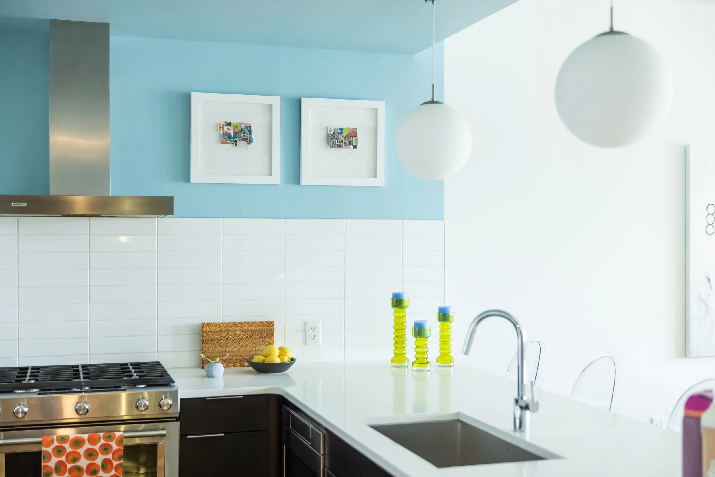 Modern kitchen with blue wall, white subway tile backsplash, white countertop, and stainless steel appliances. Fruit bowl, green candles, and abstract artwork on the wall. Pendant lights and kitchen faucet visible.