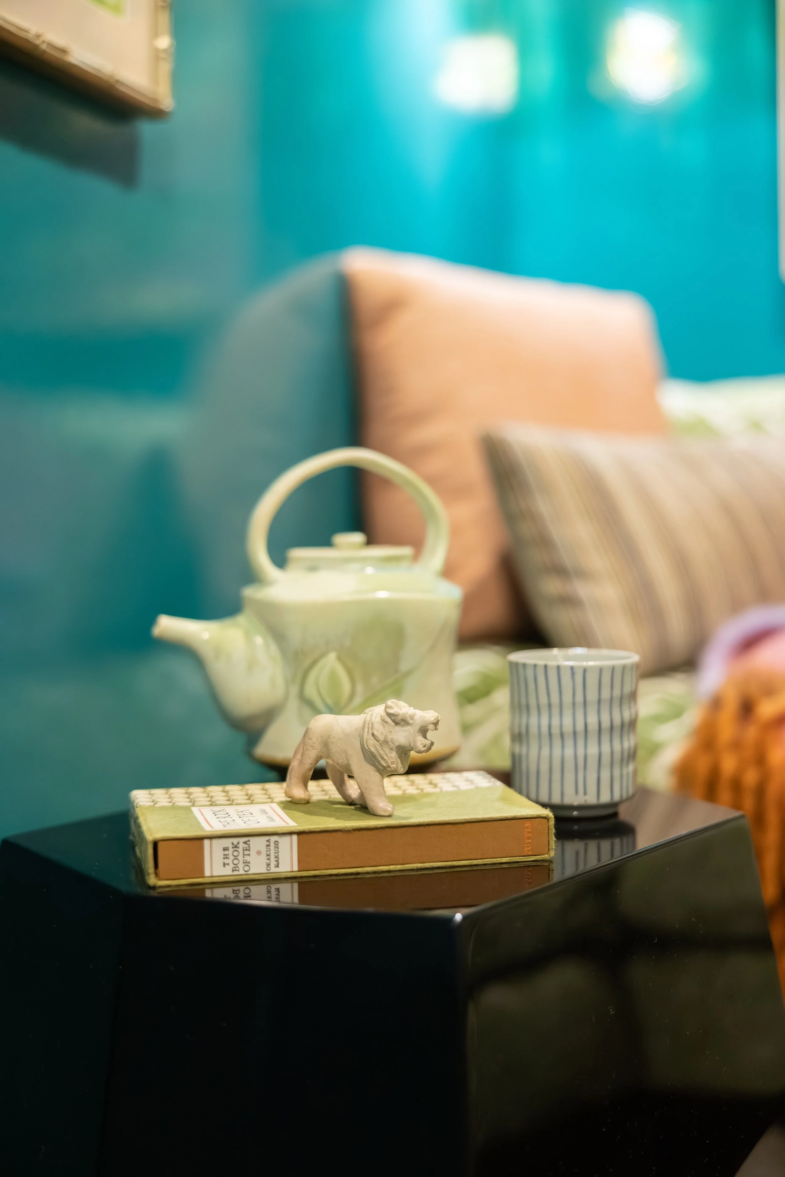 A collection of decorative items on a black surface, including a small lion figurine, a book titled 'The Book of Tea,' a green teapot, a striped mug, and pillows against a teal wall.