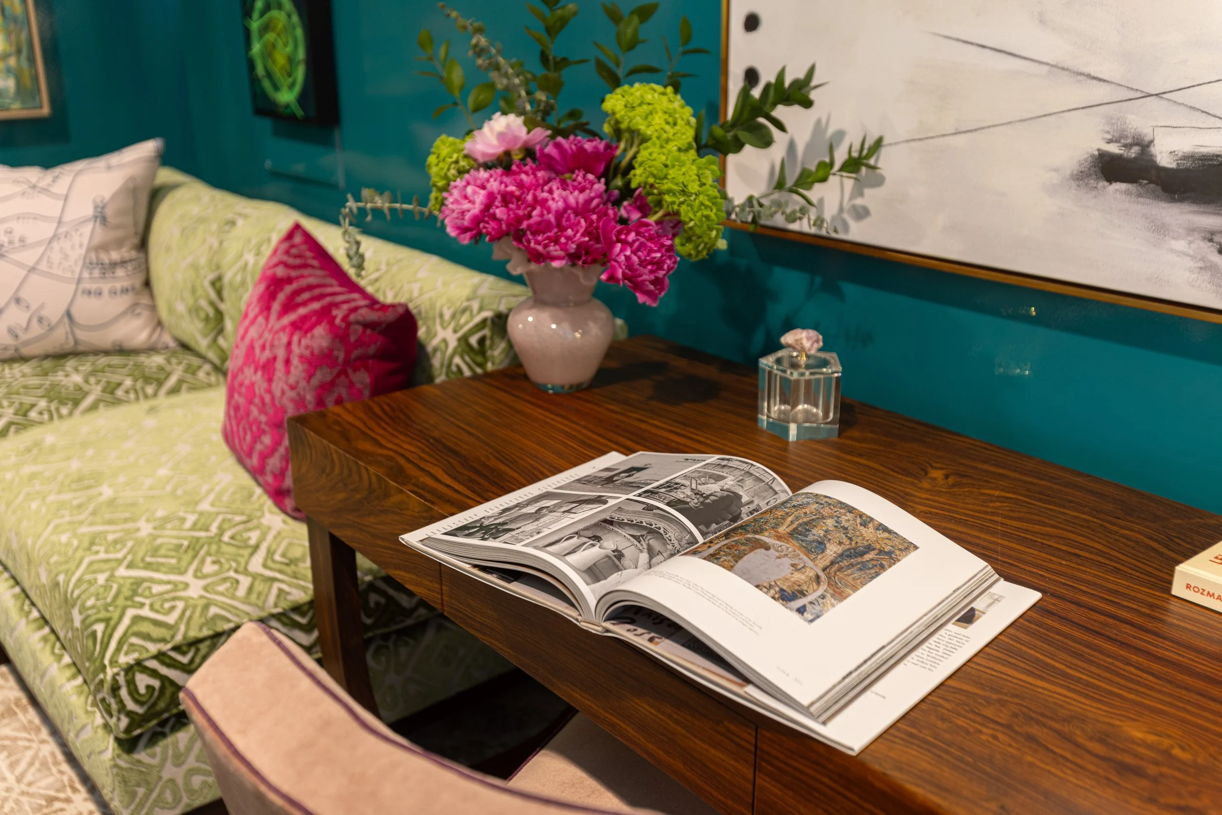 An interior scene featuring a wooden side table with an open magazine, a vase with pink and green flowers, and a small clear glass decorative object. Behind the table, there is a sofa with green patterned fabric and a pink patterned pillow, with a white and black-illustrated pillow. A teal-colored wall with framed artwork and a large abstract painting is visible in the background.