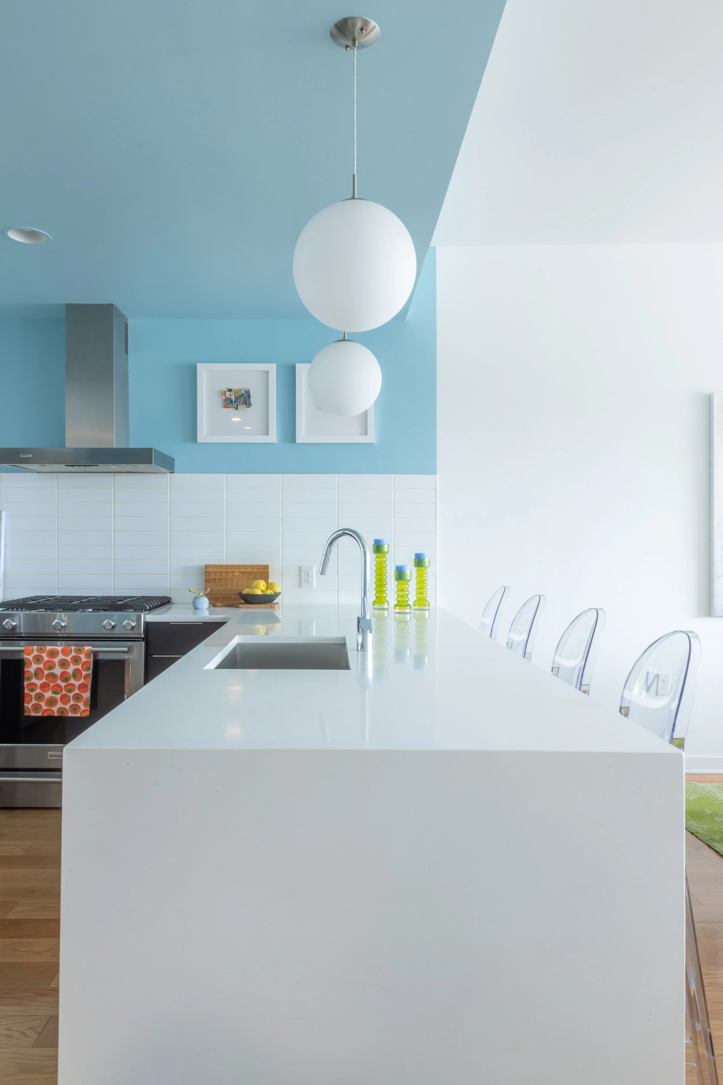 A modern kitchen with a white island, a stainless steel stove, blue and white walls, two framed pictures, a hanging light fixture, and green and yellow decorative bottles on the counter.