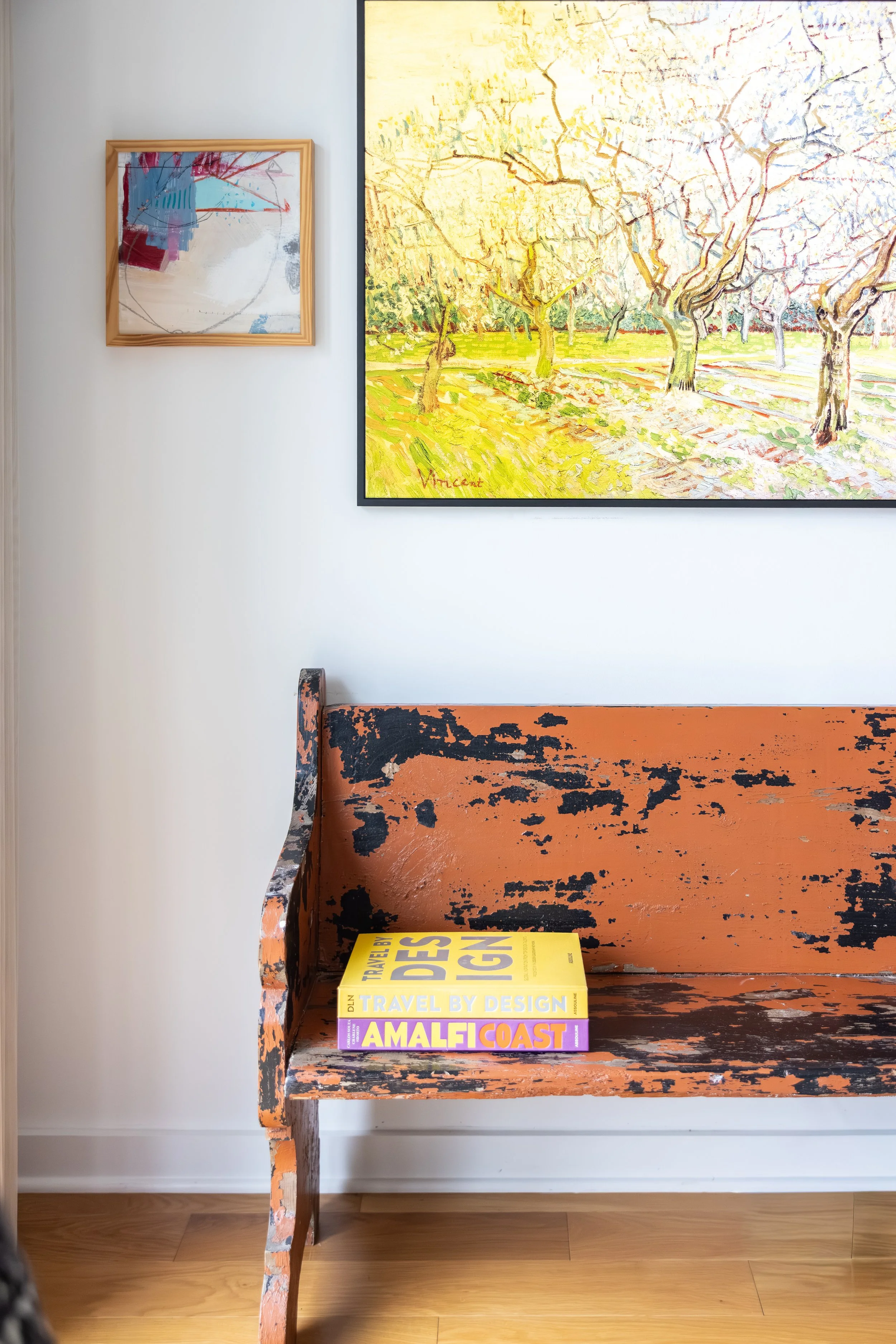 A vintage orange wooden bench with black paint chipping, placed against a white wall. On the bench, there are three travel magazines titled "Travel by Design," "Amalfi Coast," and a yellow one. Two framed paintings hang on the wall above the bench: one abstract with red, blue, and neutral tones, and another landscape with trees and grassy fields, signed "Vittore".