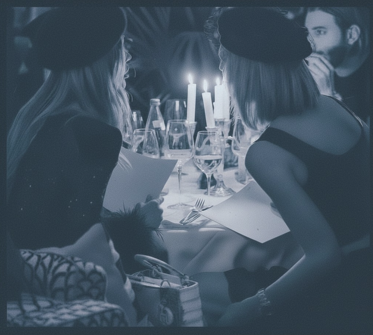 Two women sitting at a dinner table with lit candles, surrounded by glassware, bottles, and cutlery, engaging in conversation.