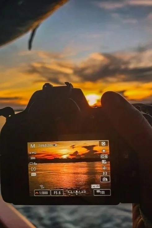 A person taking a photo of a sunset over the ocean with a digital camera from inside a vehicle.