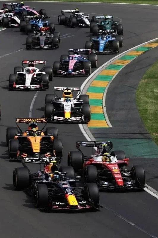 A group of Formula 1 race cars competing on a race track, with some cars close to the curb and others in a tight pack.