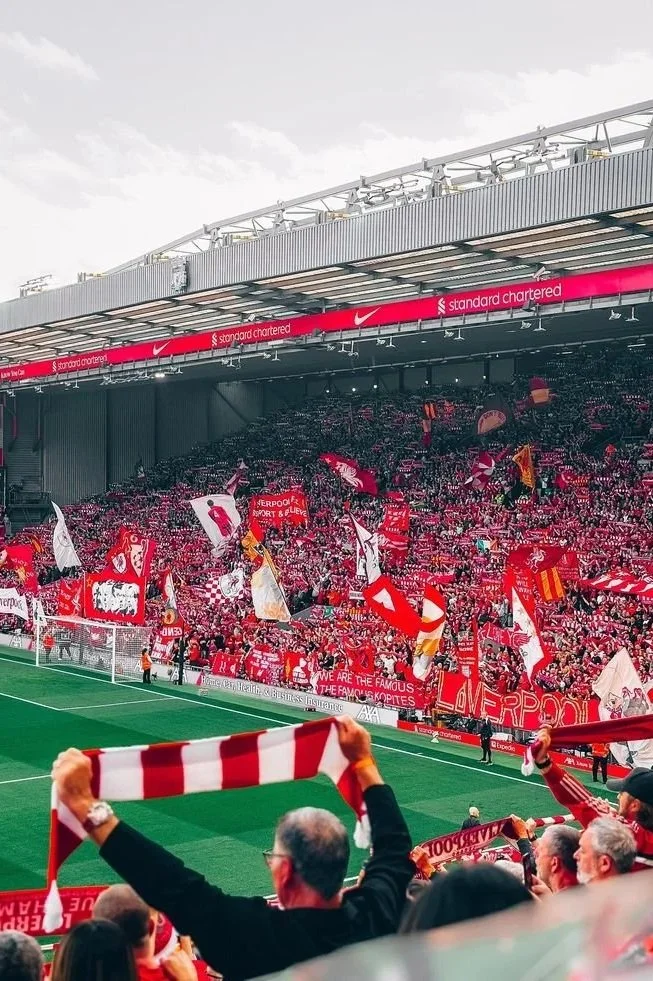 Crowd of Liverpool football fans in a stadium, waving red flags and scarves, with some standing on a green field.