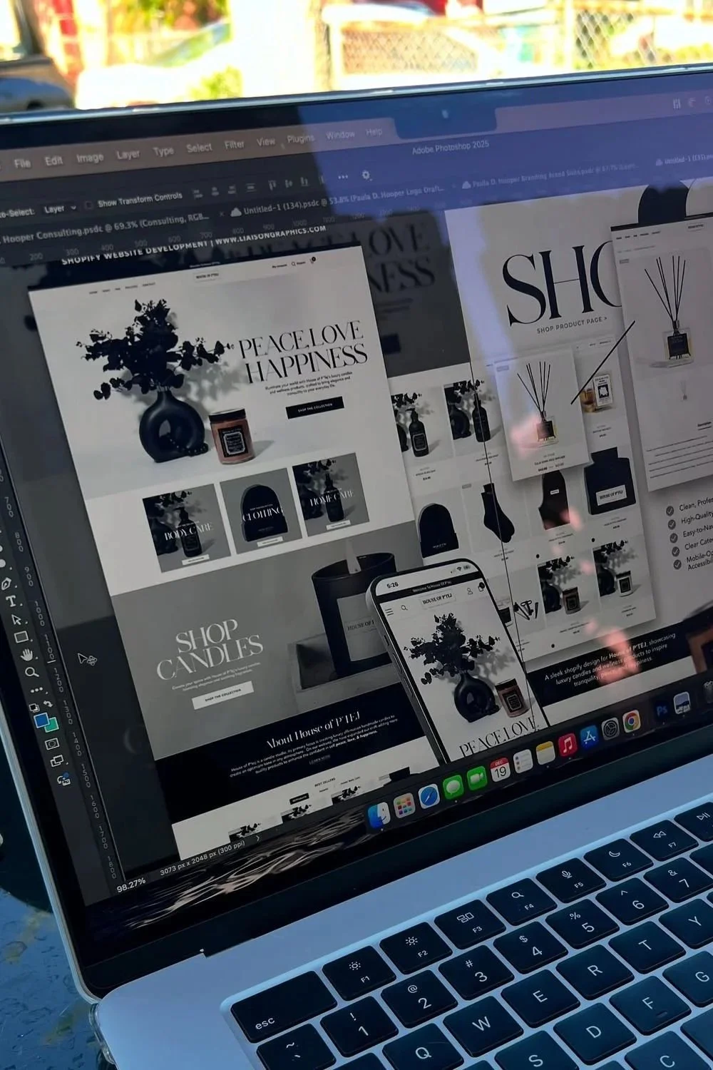 A laptop screen displays an online store webpage featuring black and white products like vases, candles, and decor items with the words 'PEACE LOVE HAPPINESS' and 'SHOP CANDLES' visible. The webpage is reflected on a smartphone screen held above, wit