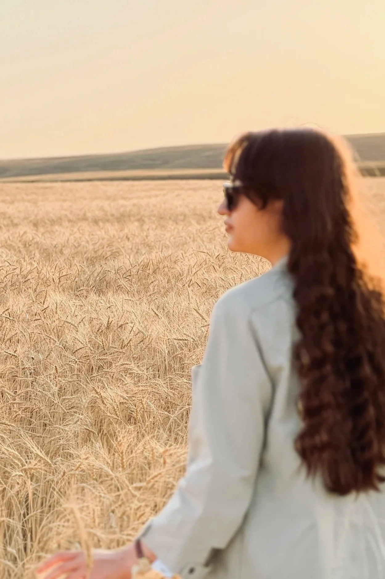 A woman with long, curly hair wearing sunglasses and a light-colored shirt walking through a wheat field at sunset.