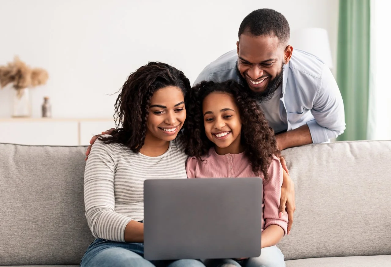 Family of three, mother, father, and daughter, sitting on a couch looking at a laptop and smiling.