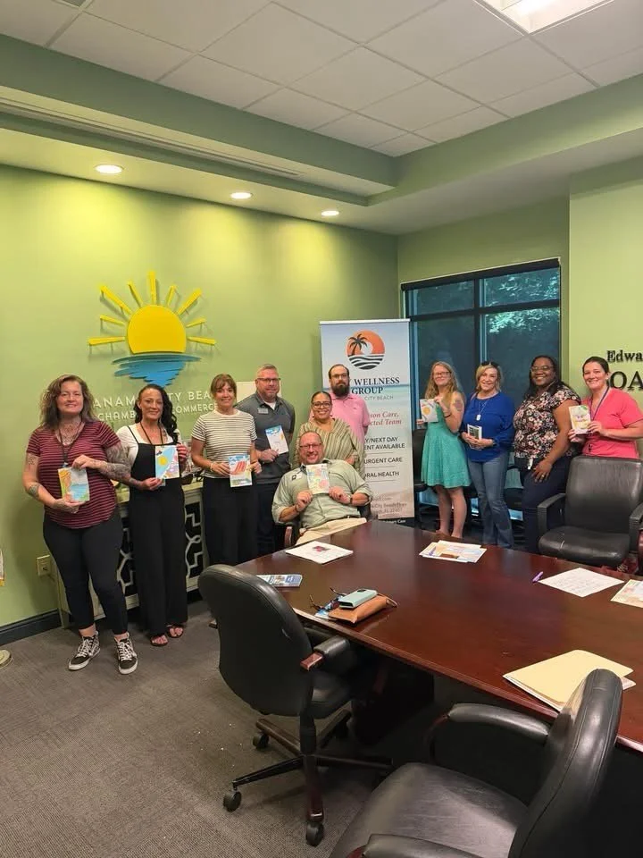 A group of eleven people posing in a conference room, some holding pamphlets, with a green wall decorated with a sun and wave logo, and a banner for Wellness Group in the background.