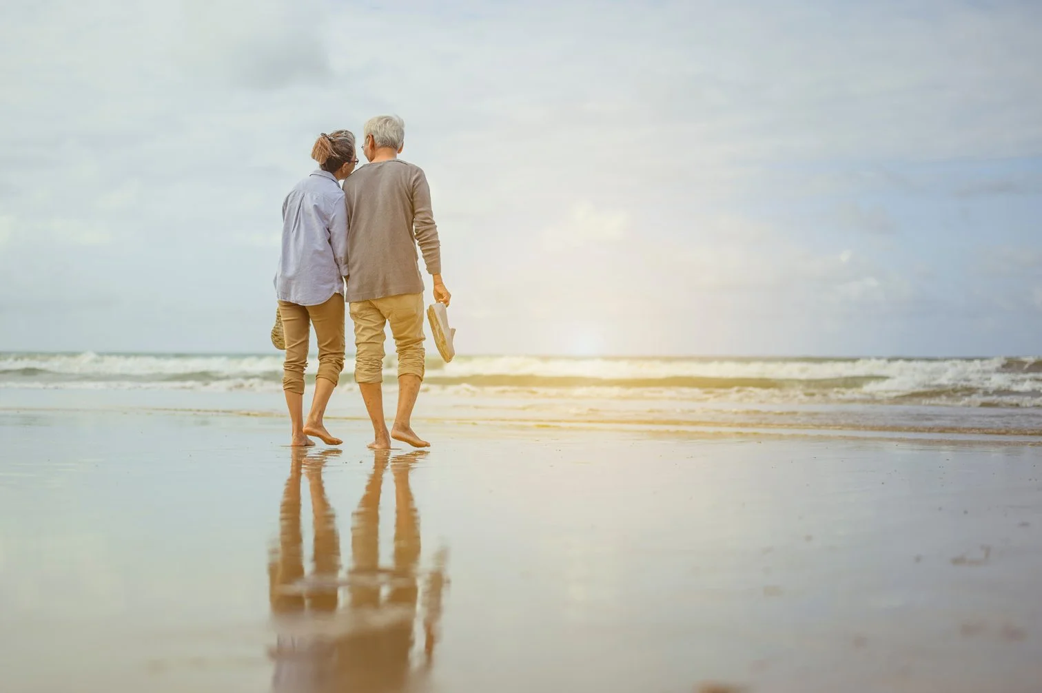 An elderly couple walking barefoot on a beach during sunset, holding hands and carrying a pair of shoes, with waves in the background.