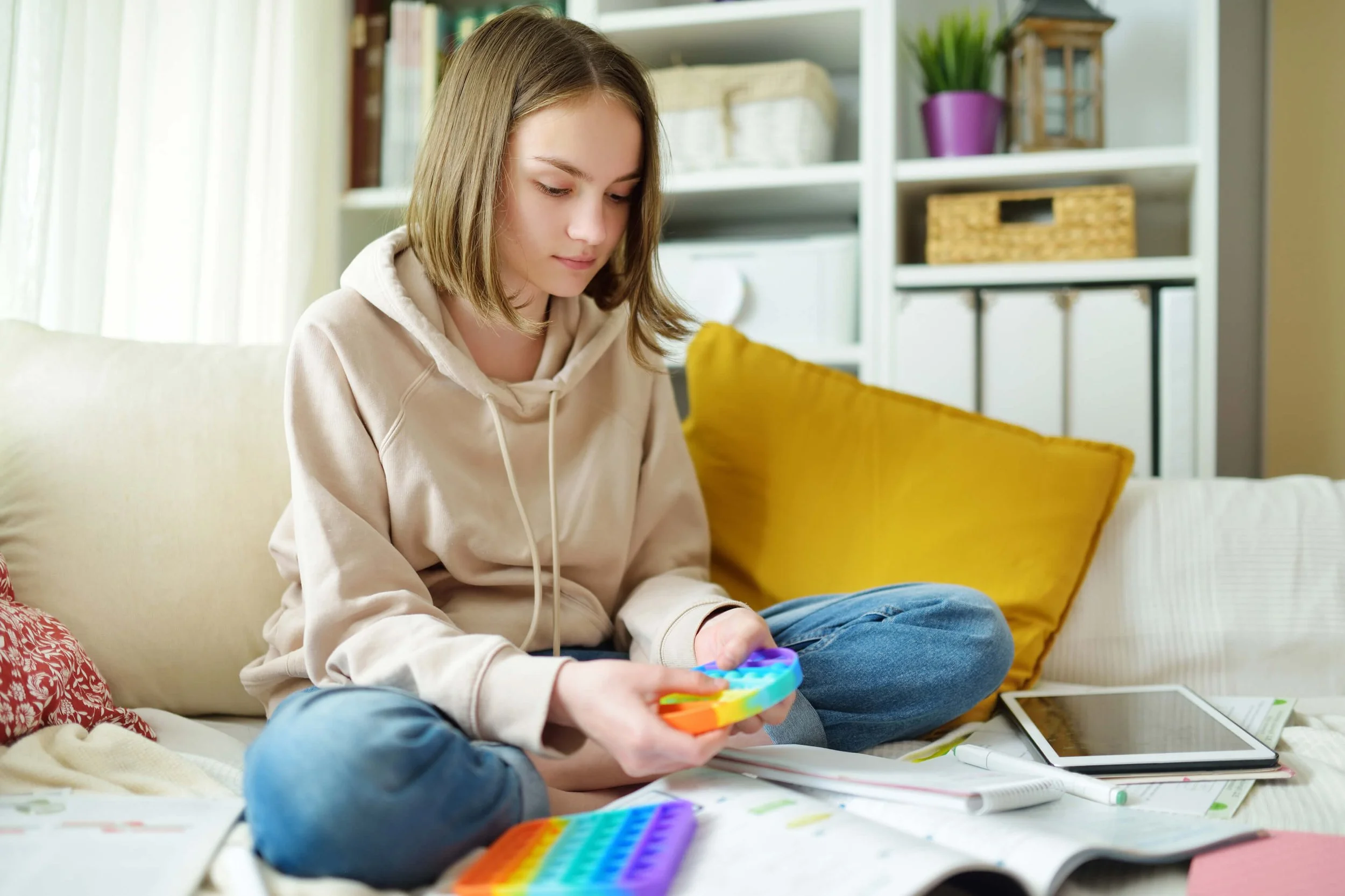 A teenage girl sitting on a beige sofa, playing with a colorful fidget spinner, surrounded by notebooks and electronic devices in a cozy, well-lit living room.