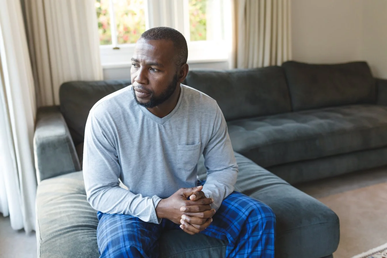 A man sitting on a gray upholstered ottoman in a living room, gazing thoughtfully out of a large window with white curtains, with a dark gray couch and a beige carpet in the background.