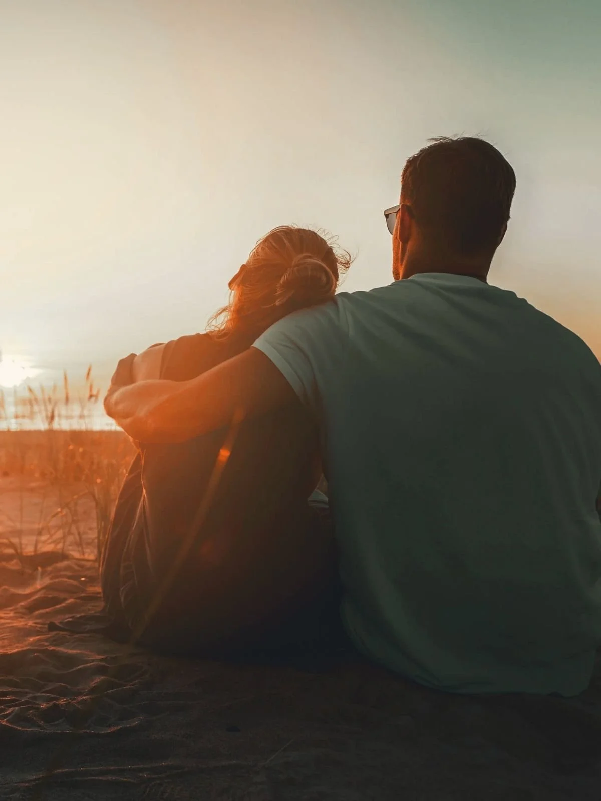 A couple sitting on the beach during sunset, with one person's arm around the other's shoulder, gazing at the horizon.