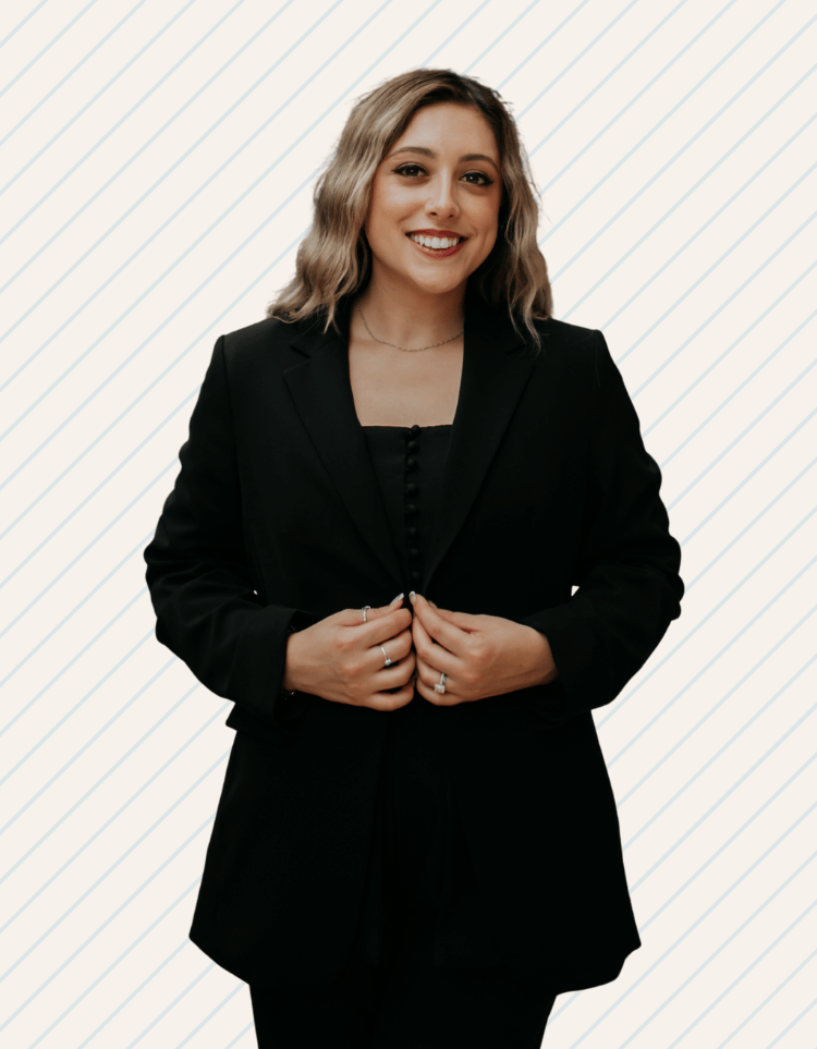 A woman with shoulder-length wavy blonde hair, wearing a black blazer and a black top, smiling and holding her hands together in front of her. Dr. Kayla Page of Bay Wellness Group.