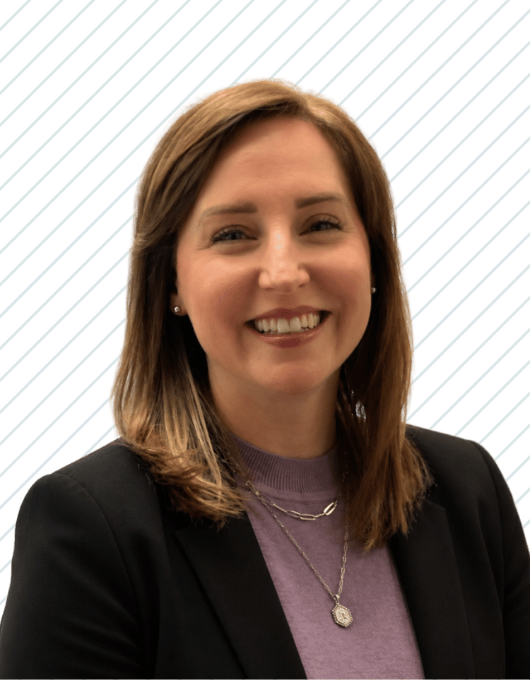 A woman with shoulder-length brown hair, smiling, wearing a black blazer, purple top, and layered necklaces, Aimee Hartzog of Bay Wellness Group.