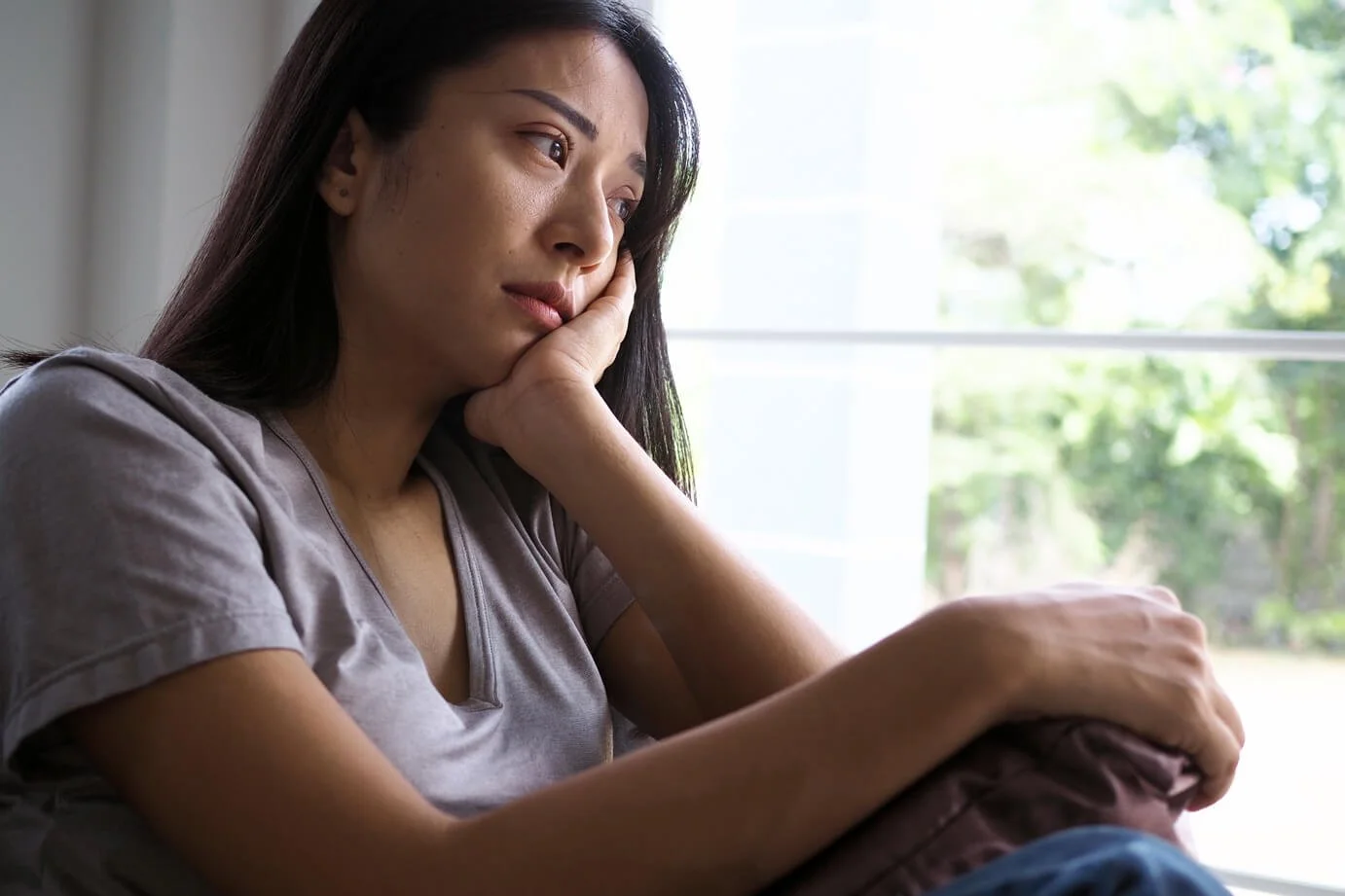 A woman sitting by a window with a thoughtful or sad expression, resting her face on her hand.