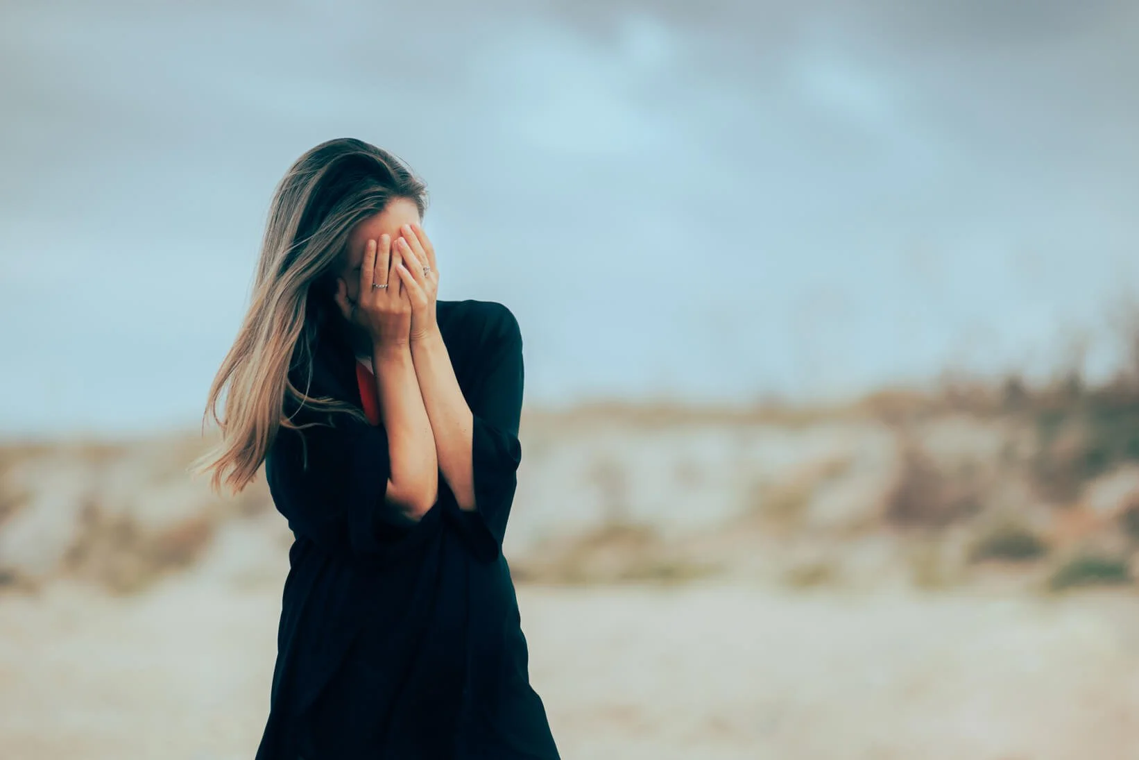 A woman standing outdoors with her hands covering her face, possibly distressed or upset, with a blurred landscape background.