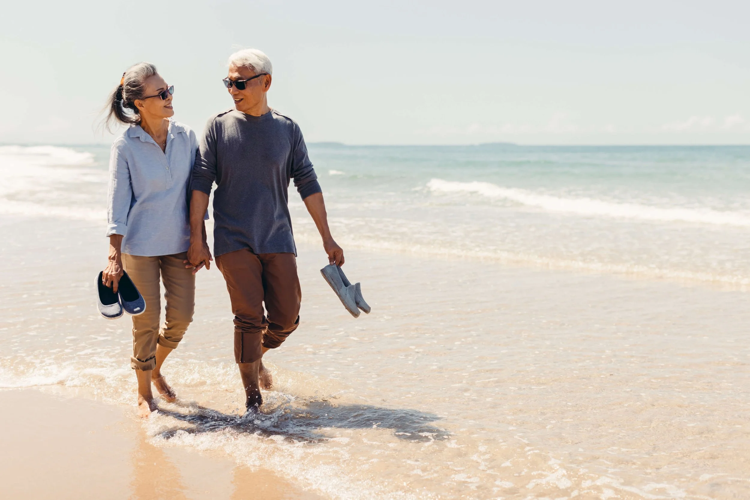 An elderly couple walking hand in hand along the beach shoreline, each holding a pair of shoes, with gentle waves and the ocean in the background.