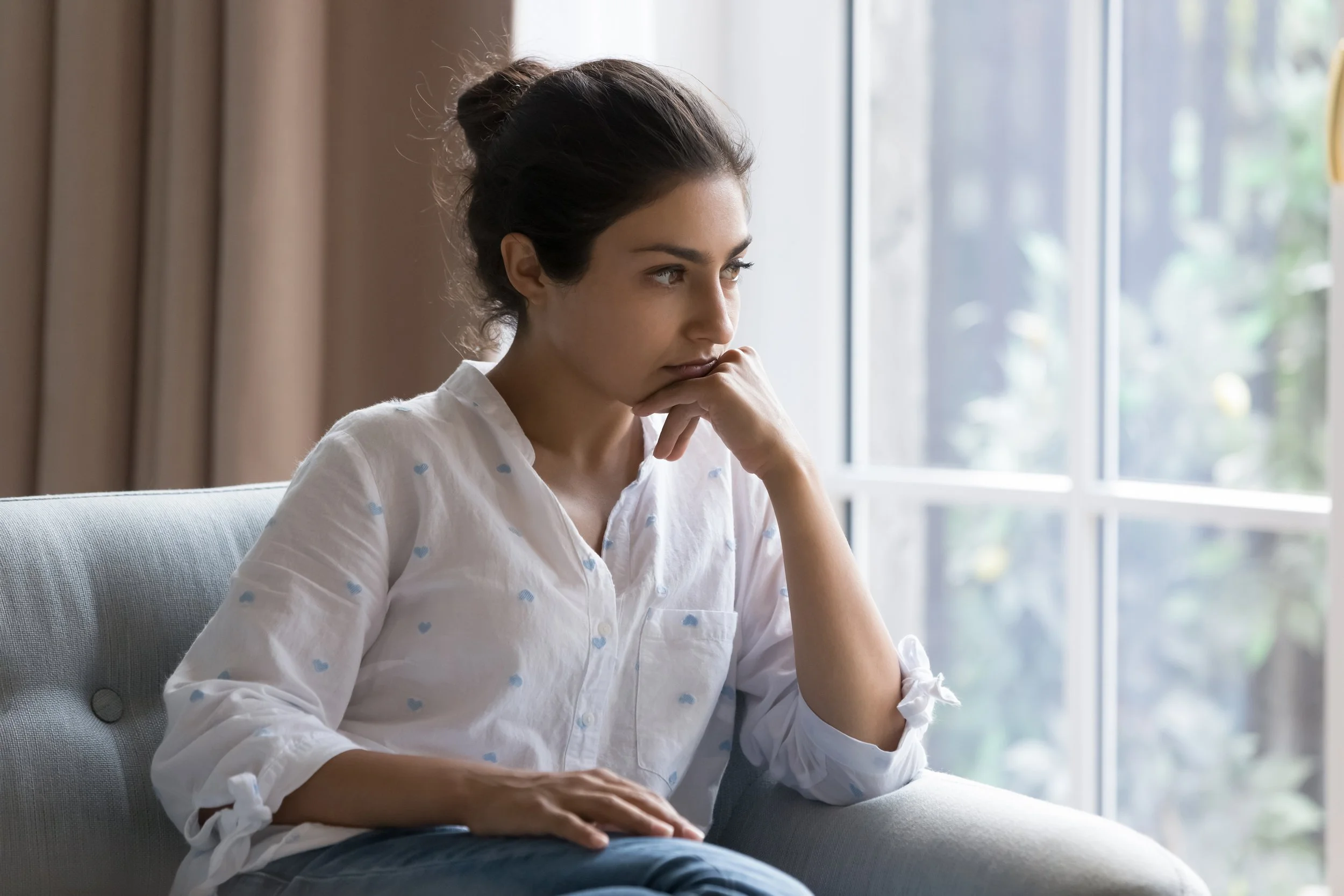 A young woman with dark hair in a bun, wearing a white blouse with blue hearts, sitting on a gray sofa near a window, looking thoughtful with her hand near her chin.