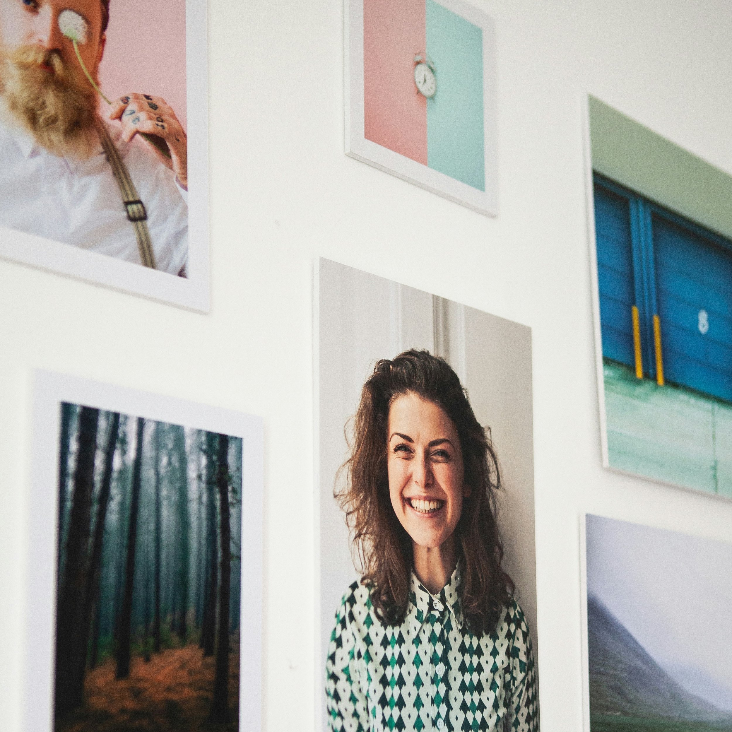Partial view of a woman with curly brown hair wearing a black and white patterned dress, smiling, standing in front of a gallery wall with nature photographs and colorful paper or canvas art.