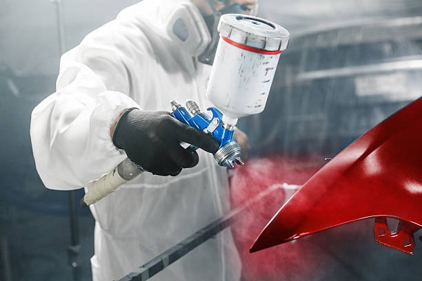 A person wearing protective gear spray painting a red car panel in an automotive shop.