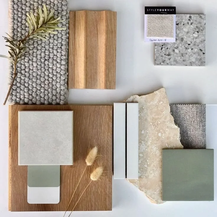 An assortment of interior design materials, including fabric swatches, wood samples, stone tiles, a sprig of greenery, and decorative elements arranged on a white surface.