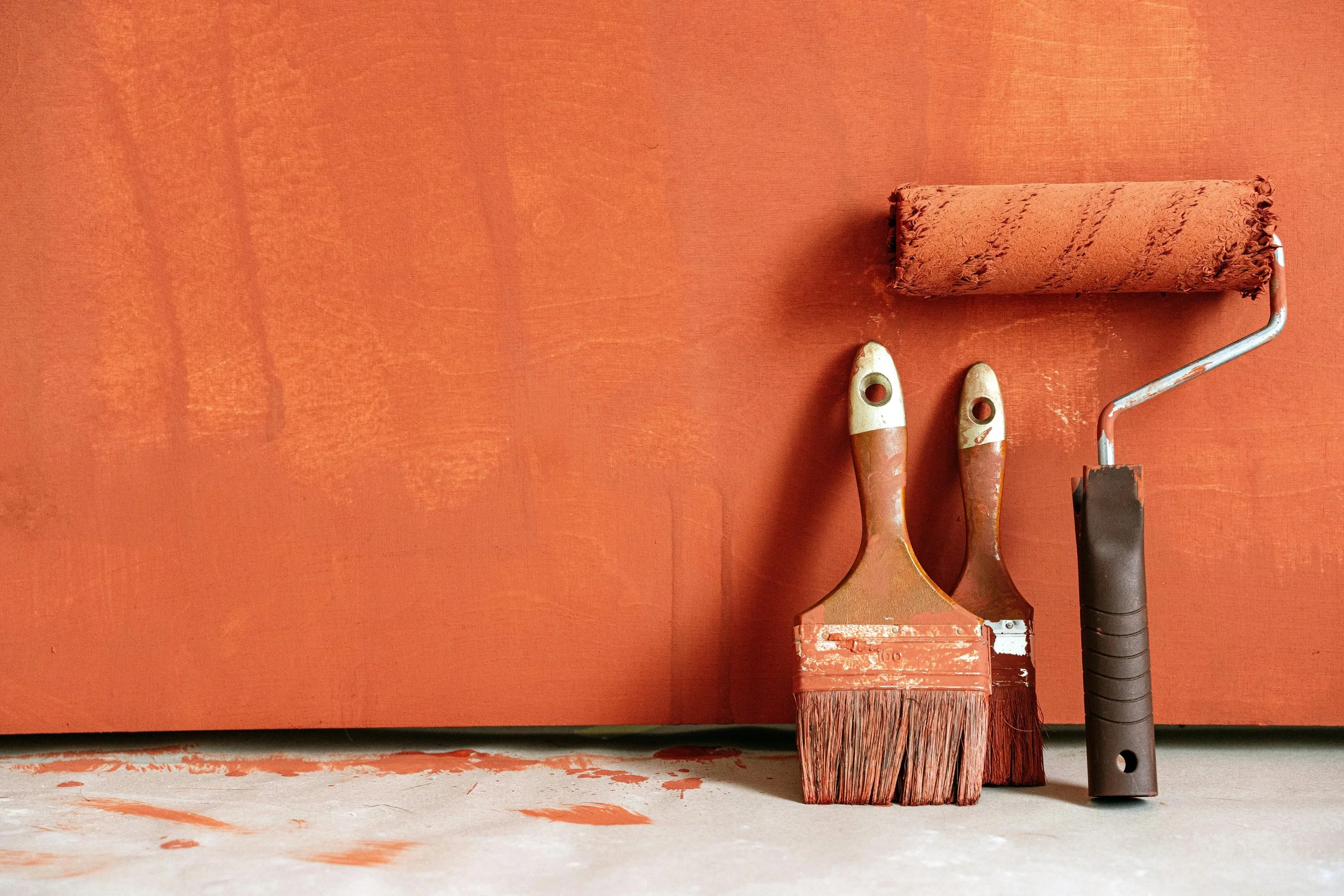Paint roller, paintbrush, and paint scraper set against a painted wall and floor, used for painting the wall in a reddish-orange color