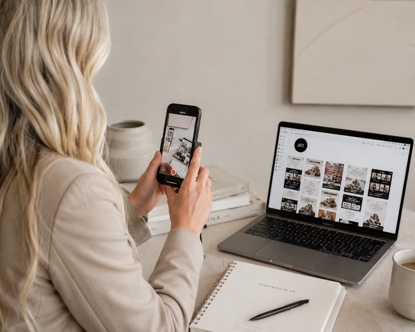 A woman with long blonde hair taking a photo of her workspace using her smartphone. On the desk, there is an open laptop displaying a social media or marketing page, a spiral notebook titled 'Content Plan' with a pen, a cup of coffee, and some stacked books.