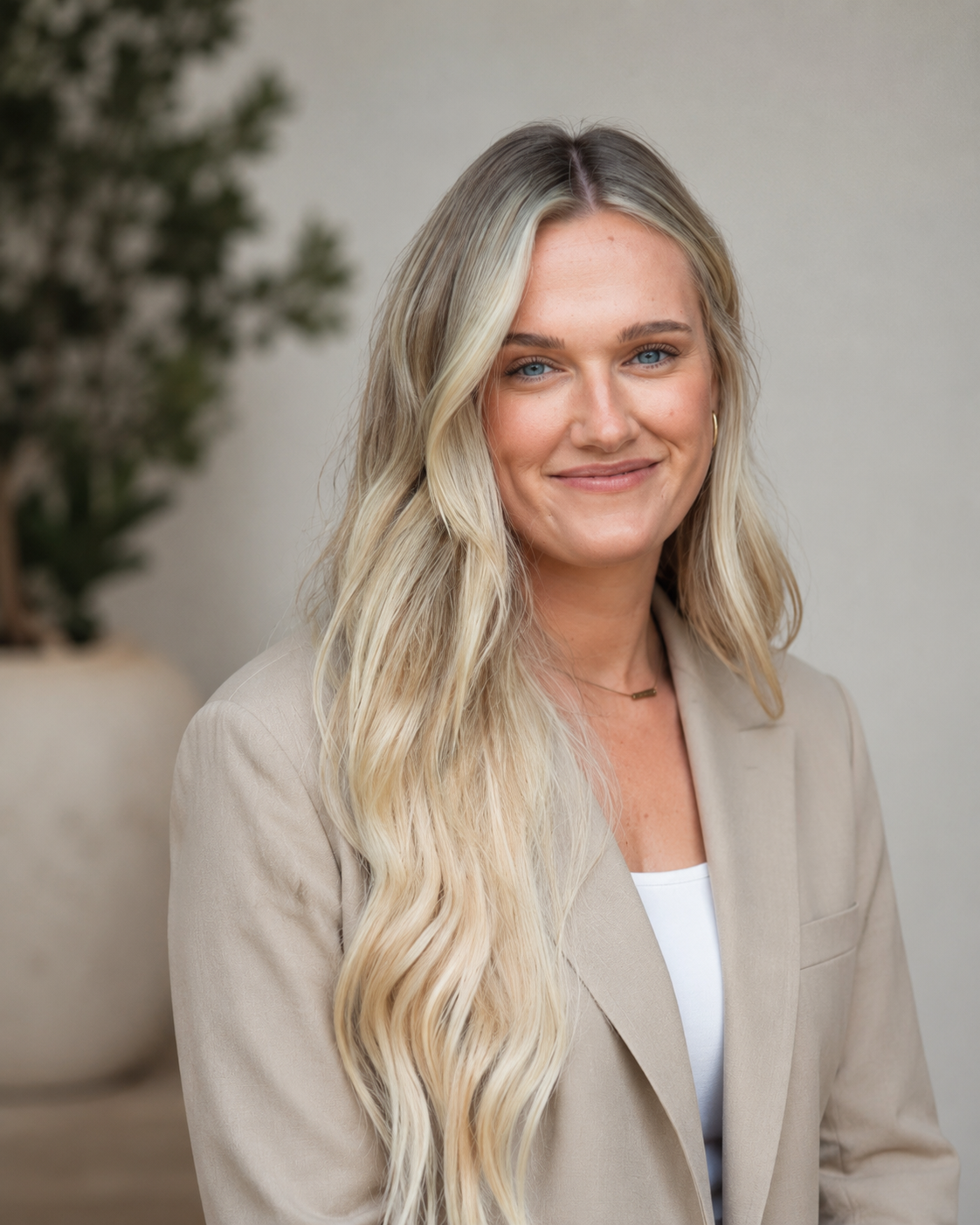 A woman with long blonde wavy hair, wearing a beige blazer and white top, smiling while standing indoors with a blurred background of a plant and neutral-colored wall.