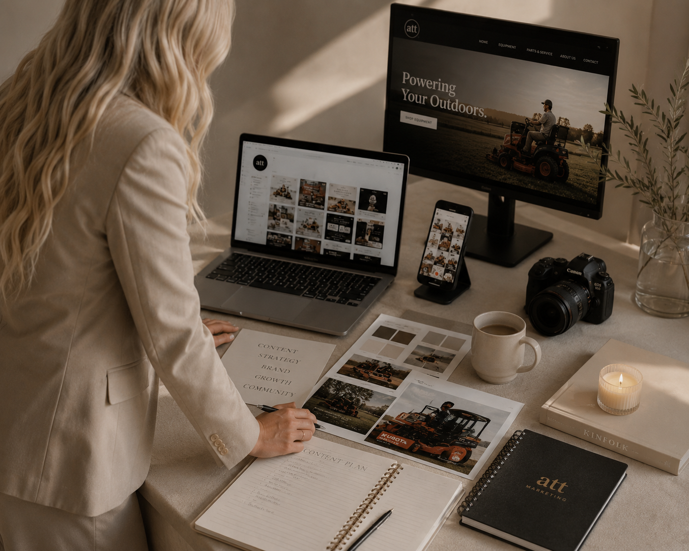 A woman with long blonde hair wearing a beige blazer working at a cluttered desk. The desk has a laptop, a monitor displaying outdoor equipment website, a smartphone, a Canon camera, a cup of coffee, a lit candle, a book titled "Kinfolk," a black notebook labeled "att marketing," a spiral notepad, and printed photographs of outdoor machinery and landscapes. The woman appears to be planning content with printed photos, a content plan, and a marketing notebook.