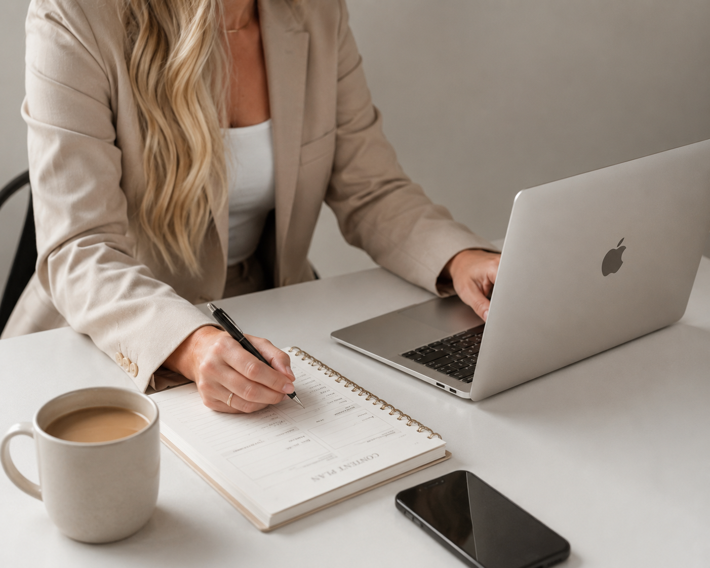 A woman in a beige jacket working at a white desk with a laptop, coffee mug, smartphone, and a notebook.