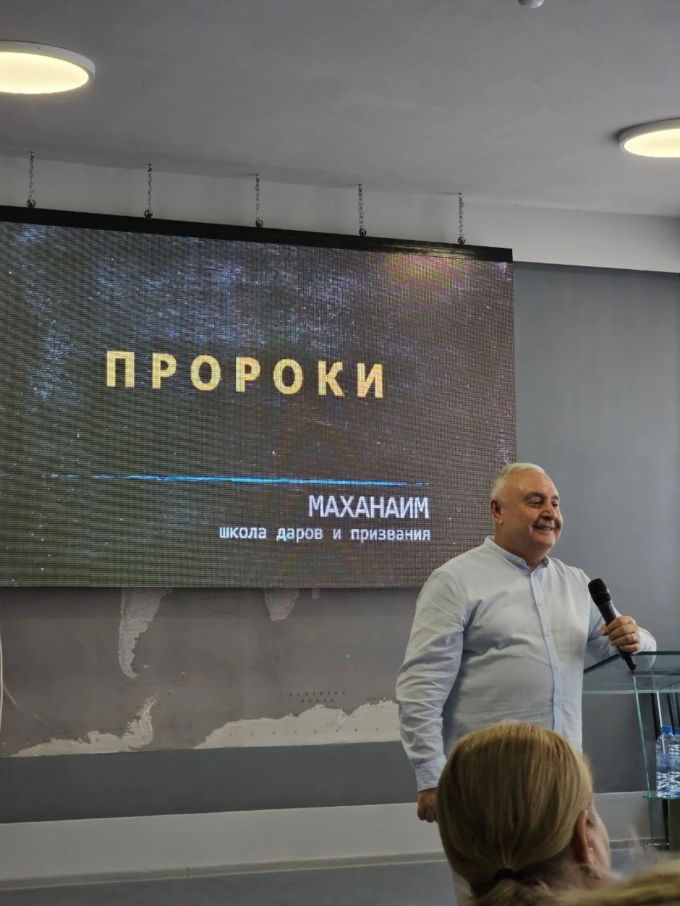 Տղամարդը միկրոֆոնով խոսում է ներկայացման ժամանակ, իսկ նրա ետևում մեծ էկրան է, որը ցուցադրում է ռուսերեն տեքստ: