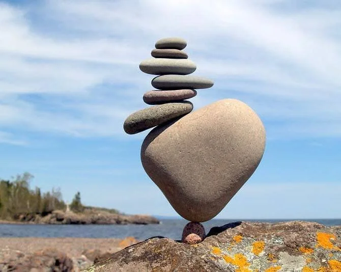Stacked stones balanced on a larger rock near the beach with a cloudy sky and distant shoreline in the background.