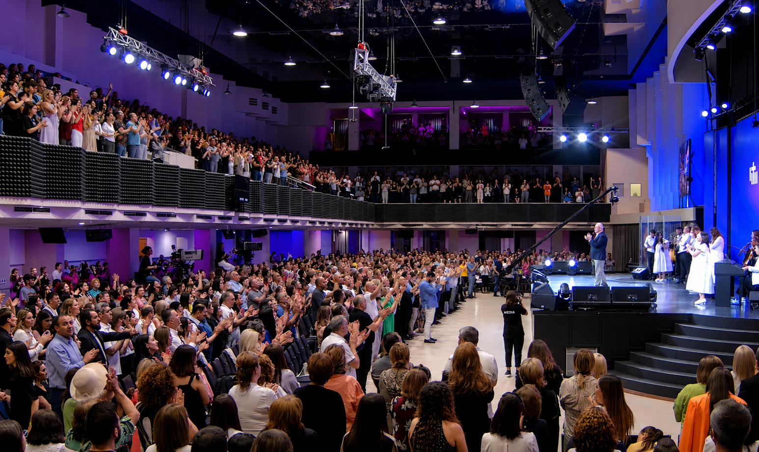 Crowd of people inside a large auditorium, standing and clapping, facing a stage where a speaker is addressing them, with a choir or group of performers on stage to the right.