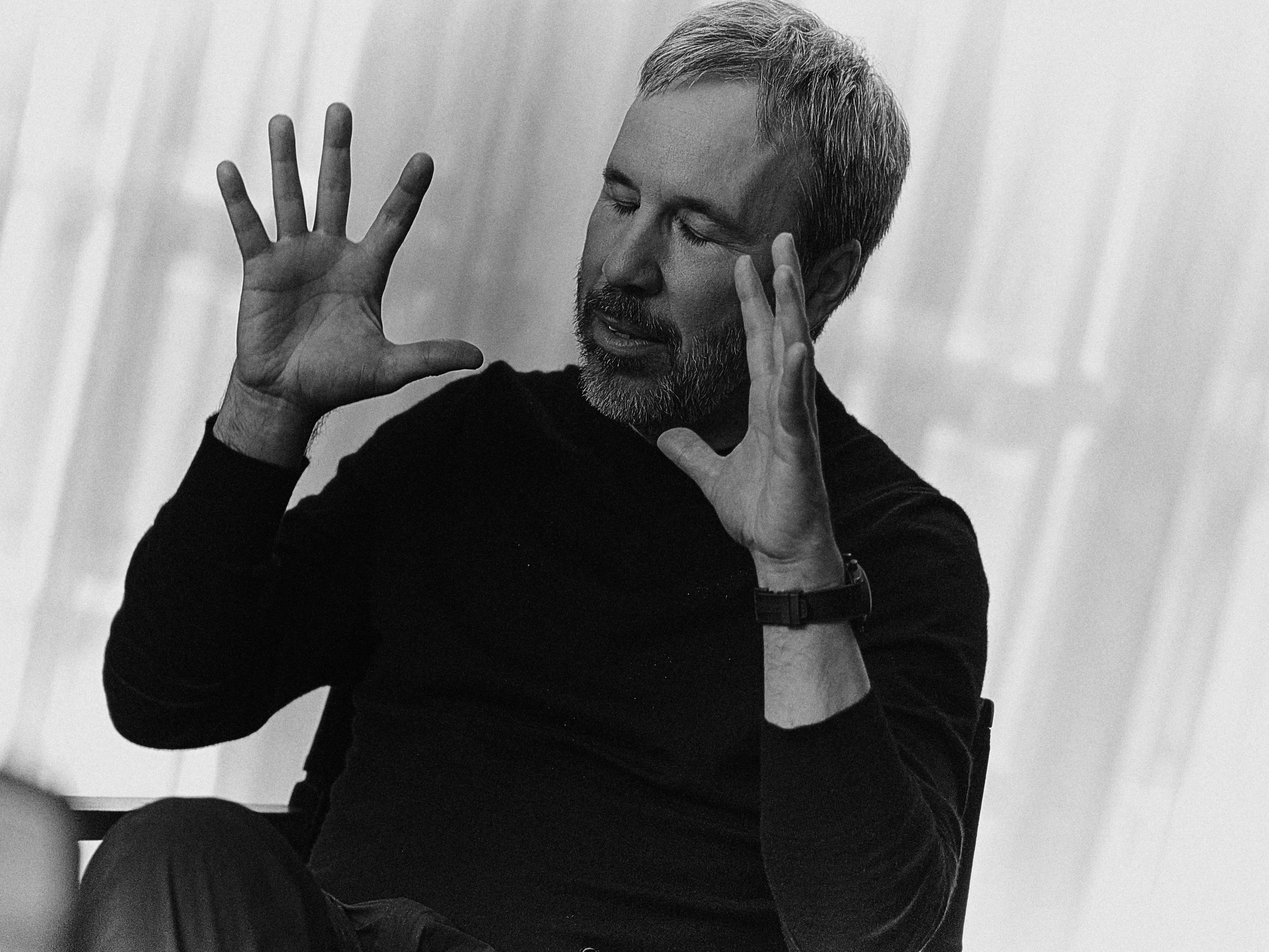 A man with short hair and a beard is sitting with his eyes closed, touching his face with both hands, against a neutral background.
