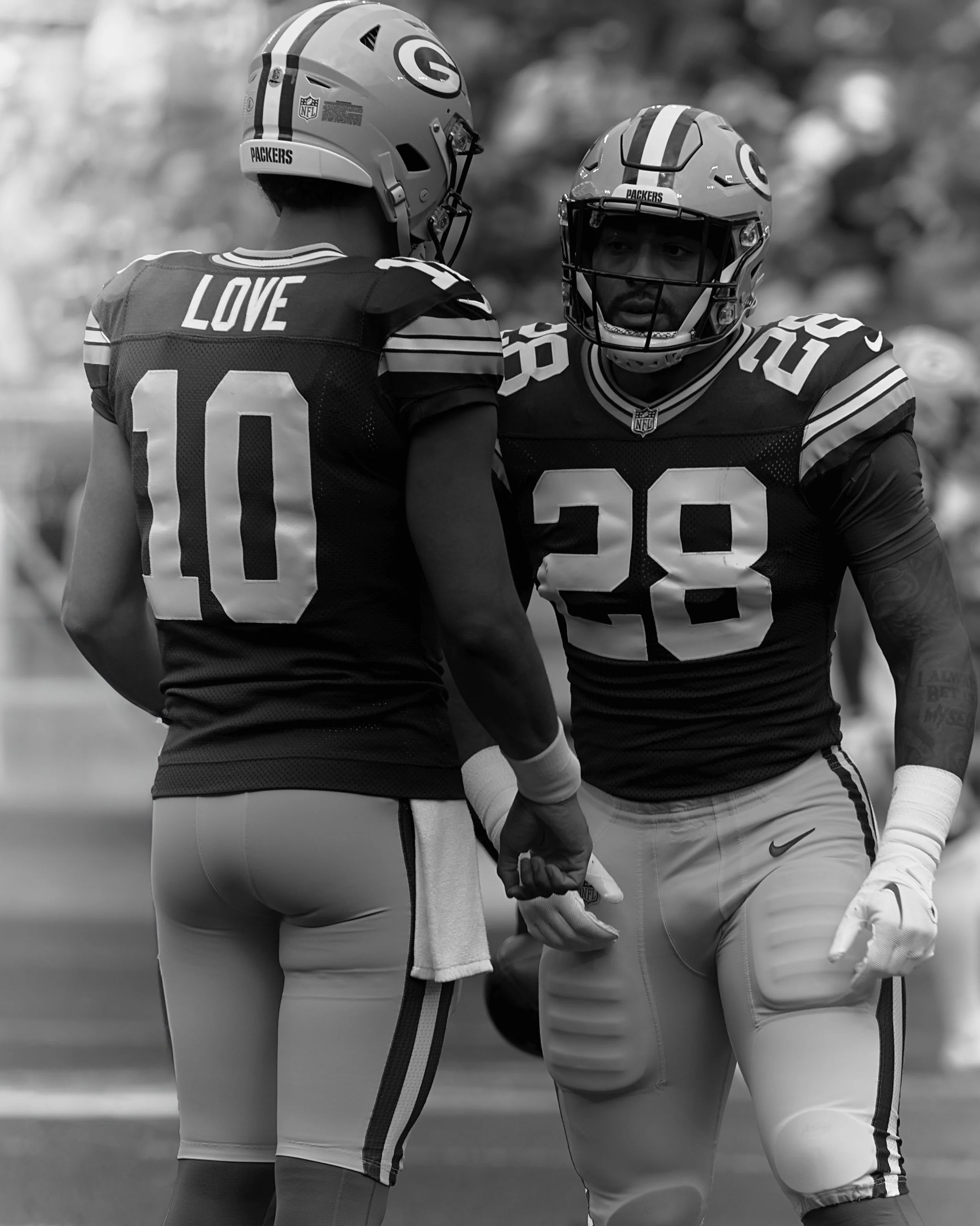Two American football players from the Green Bay Packers are standing close together, wearing helmets and jerseys, on the field during a game. One player has the name 'LOVE' and the number 10, and the other has the number 20.