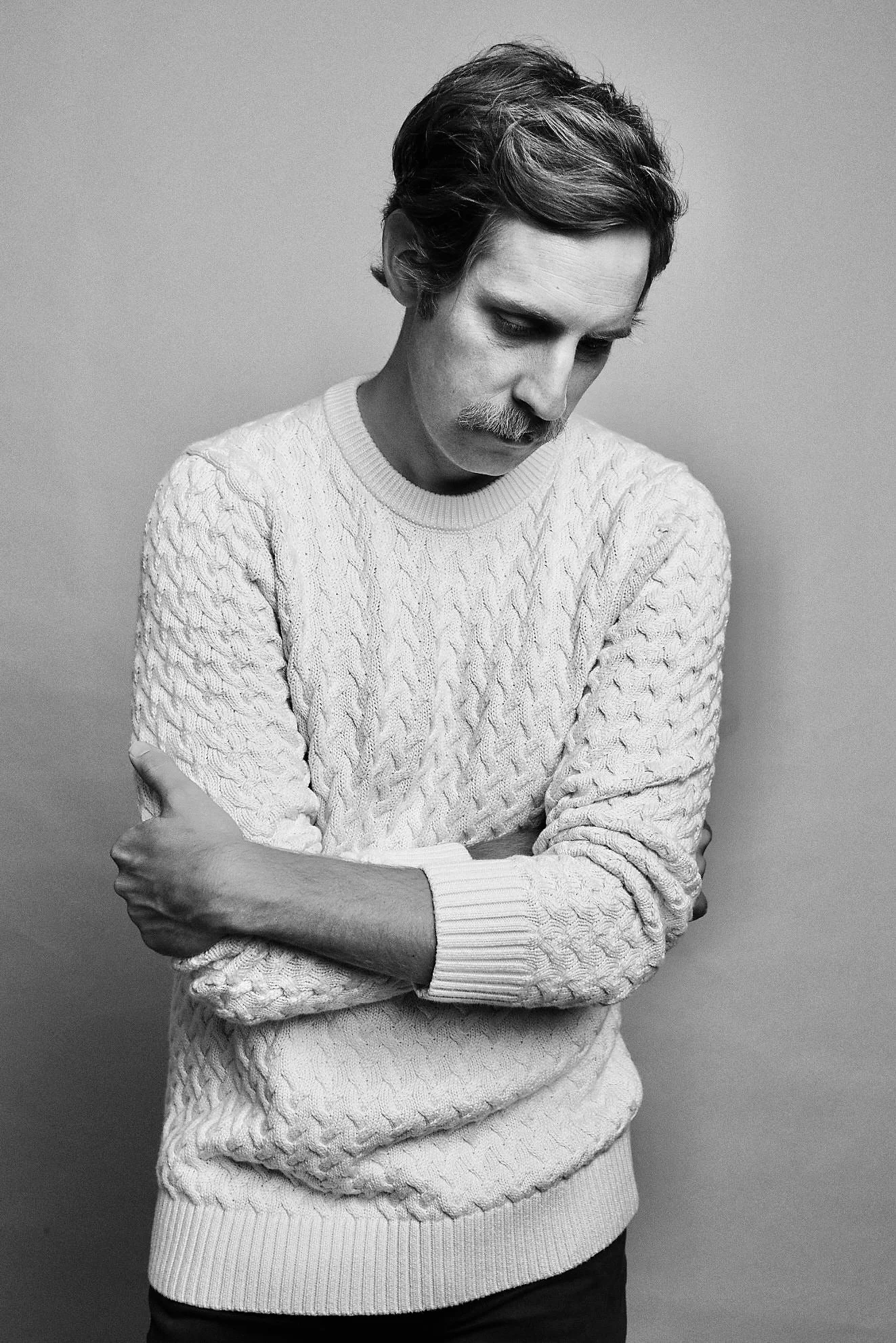 A black and white photo of a man with dark hair, a mustache, and a beard, wearing a textured cable-knit sweater, looking down with his arms crossed.