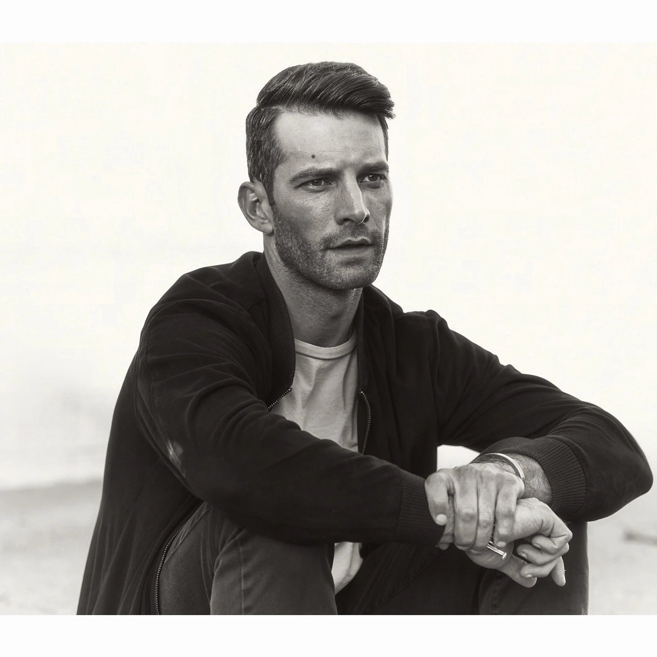 Black and white photo of a man with short, combed hair and slight stubble, wearing a dark jacket and light shirt, sitting with his arms crossed, against a plain background.