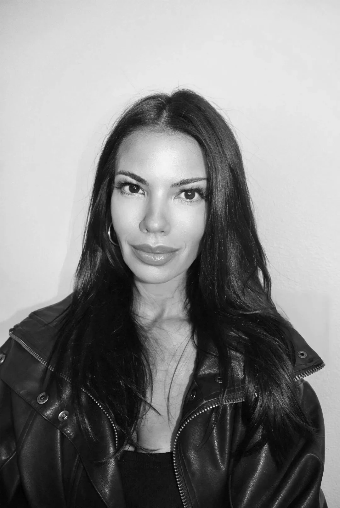 Black and white portrait of Angela Forte with long dark hair, wearing a leather jacket and hoop earrings, standing against a plain wall.