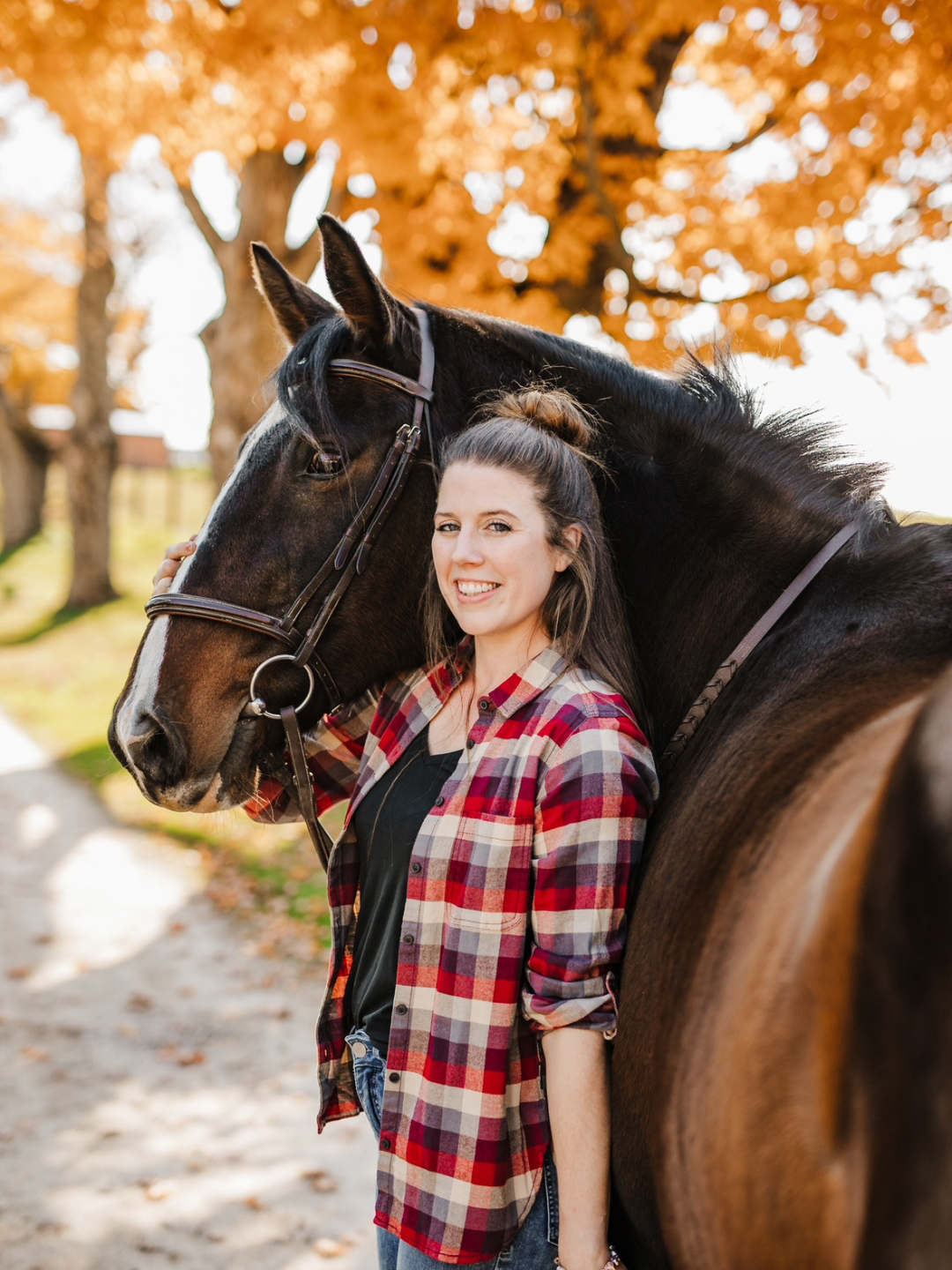 A woman in a plaid shirt standing beside a horse with autumn trees in the background.