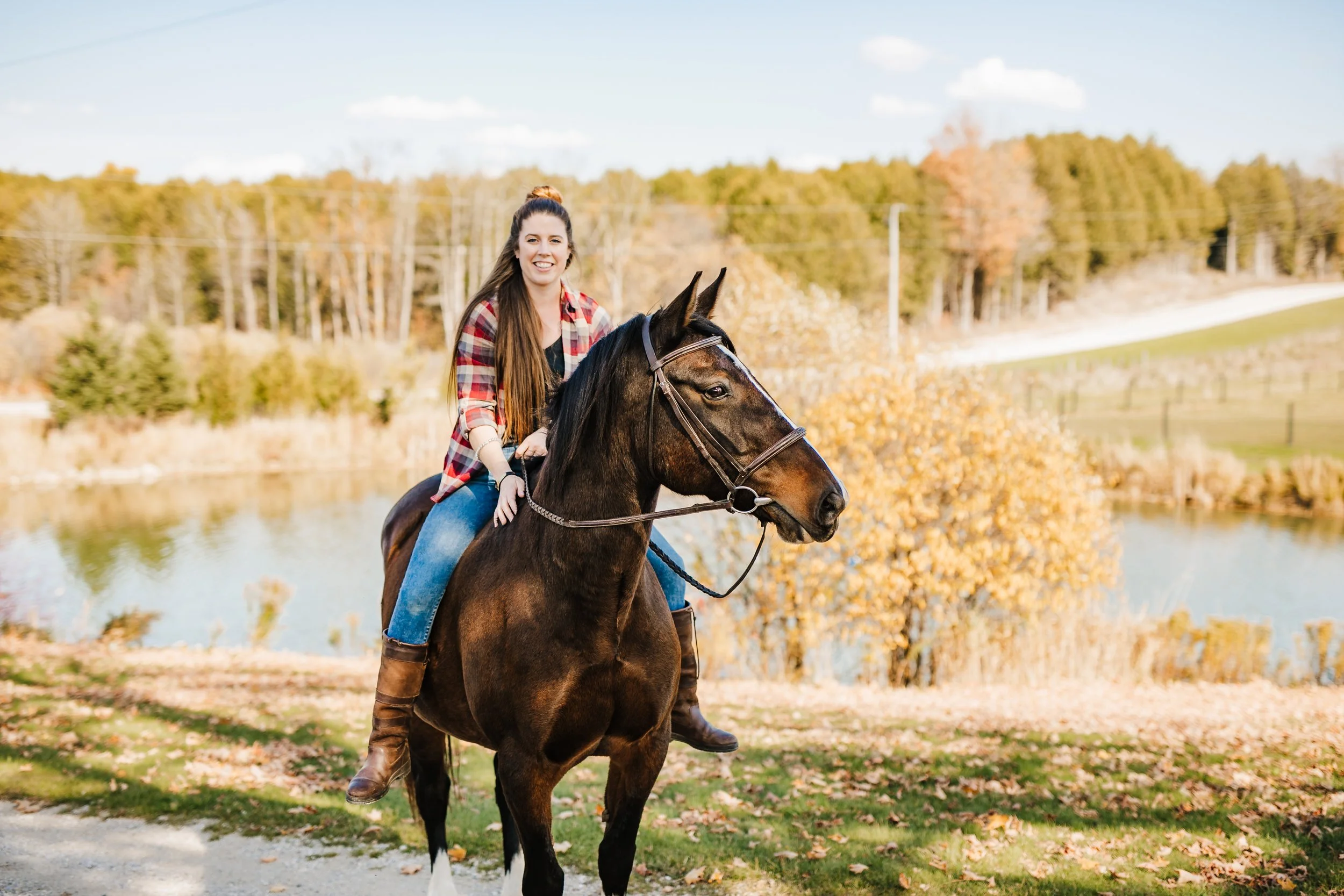 A woman with long brown hair, wearing a red plaid shirt, riding a dark brown horse with a white star on its forehead near a body of water. Fall foliage and trees are in the background.