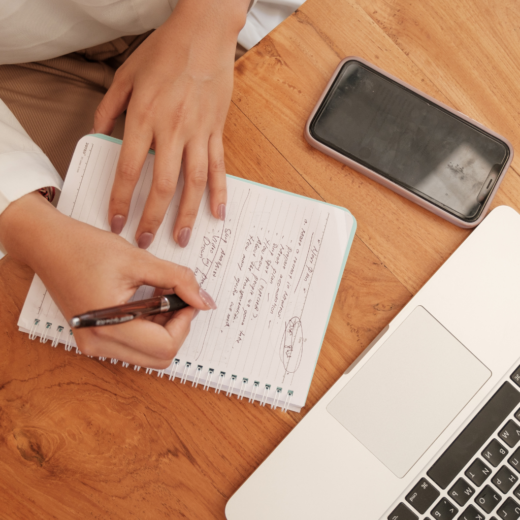 Person writing in a notebook with a pen on a wooden desk, next to a smartphone and a laptop.