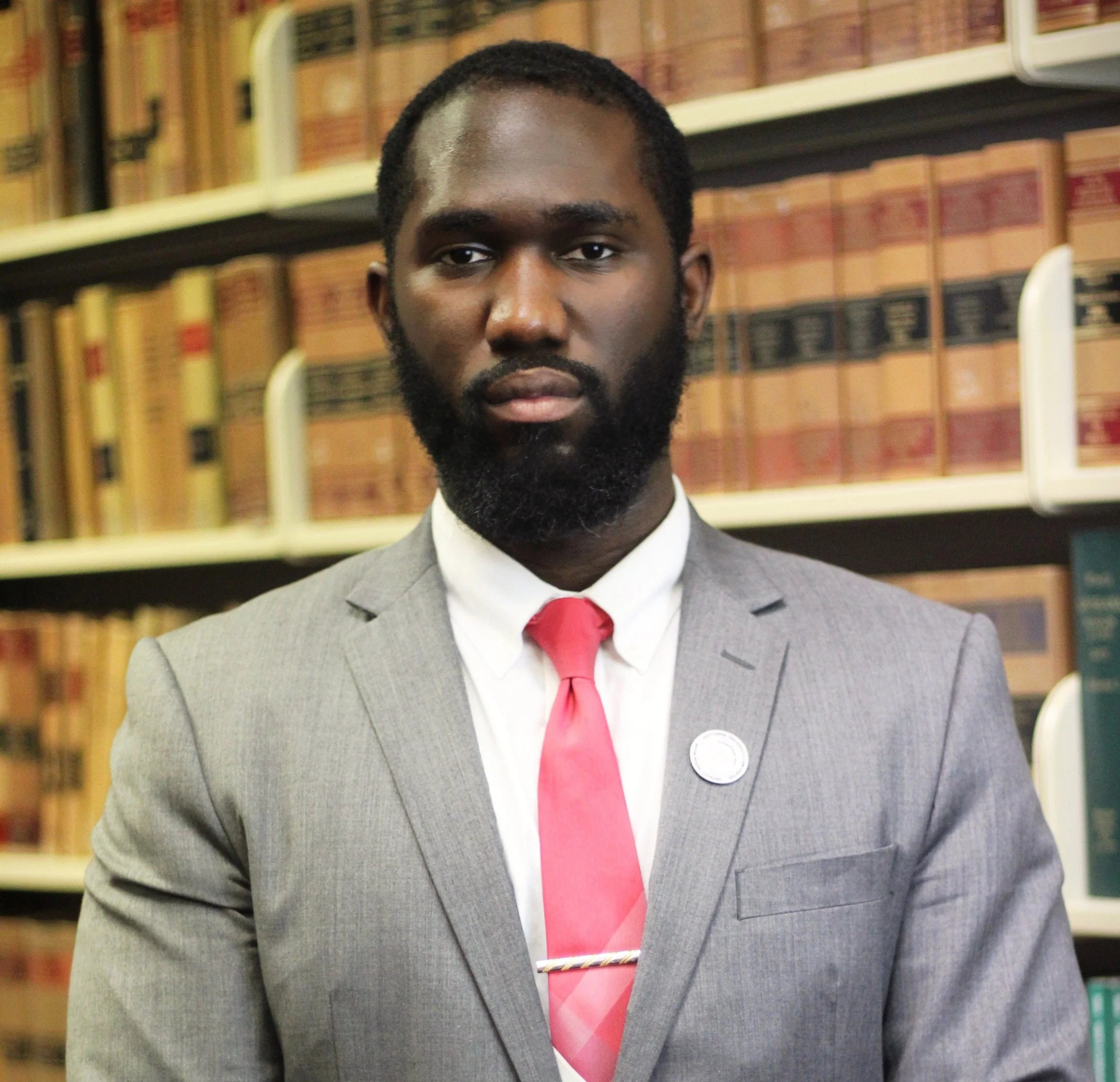 A portrait of a Black man with a beard, wearing a light gray suit and a red tie, standing in front of bookshelves filled with law books.