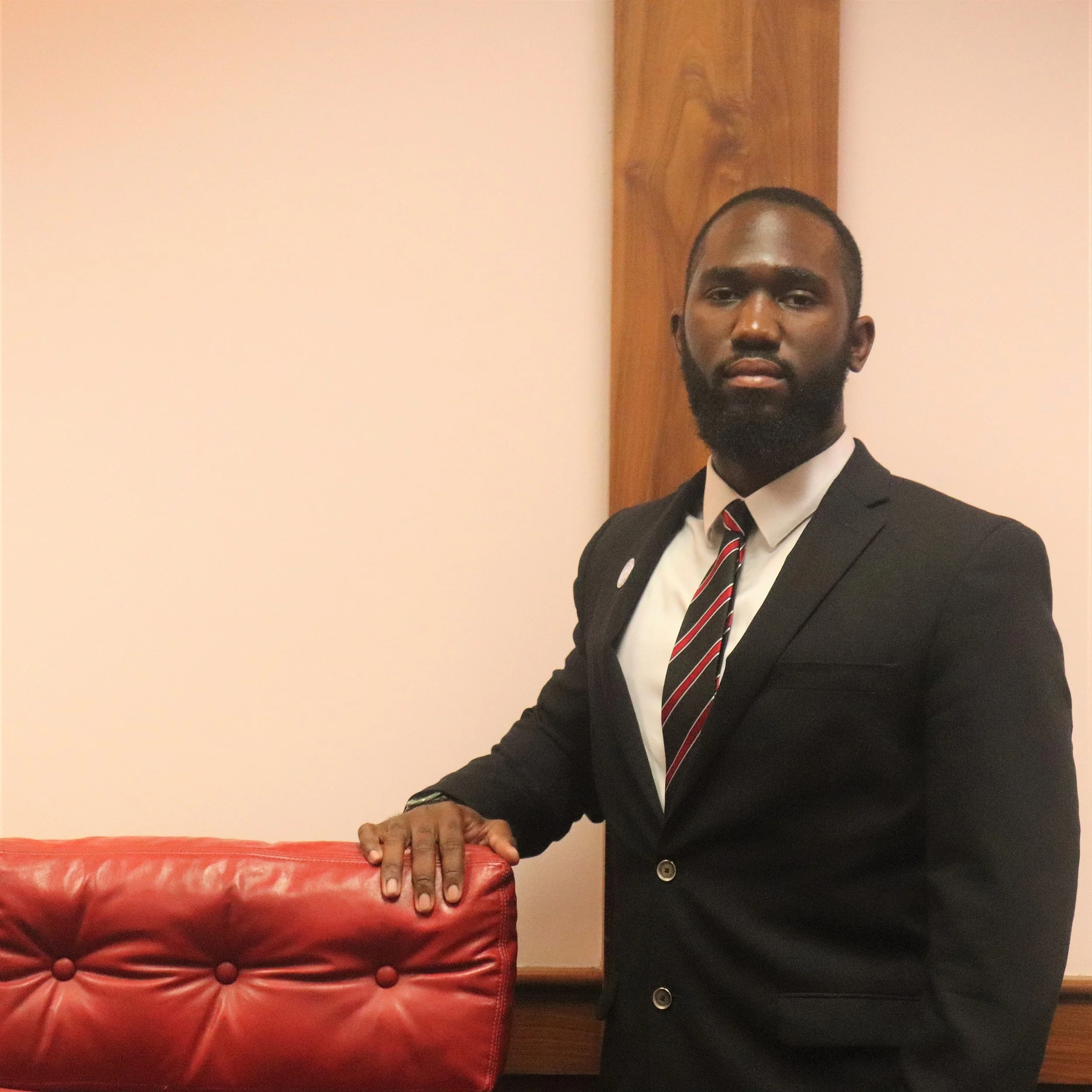 A man with dark skin, a beard, and short hair, wearing a black suit, a white shirt, and a striped tie, is standing beside a red upholstered piece of furniture with buttons. He is looking at the camera in a room with beige walls and a wooden beam in the background.