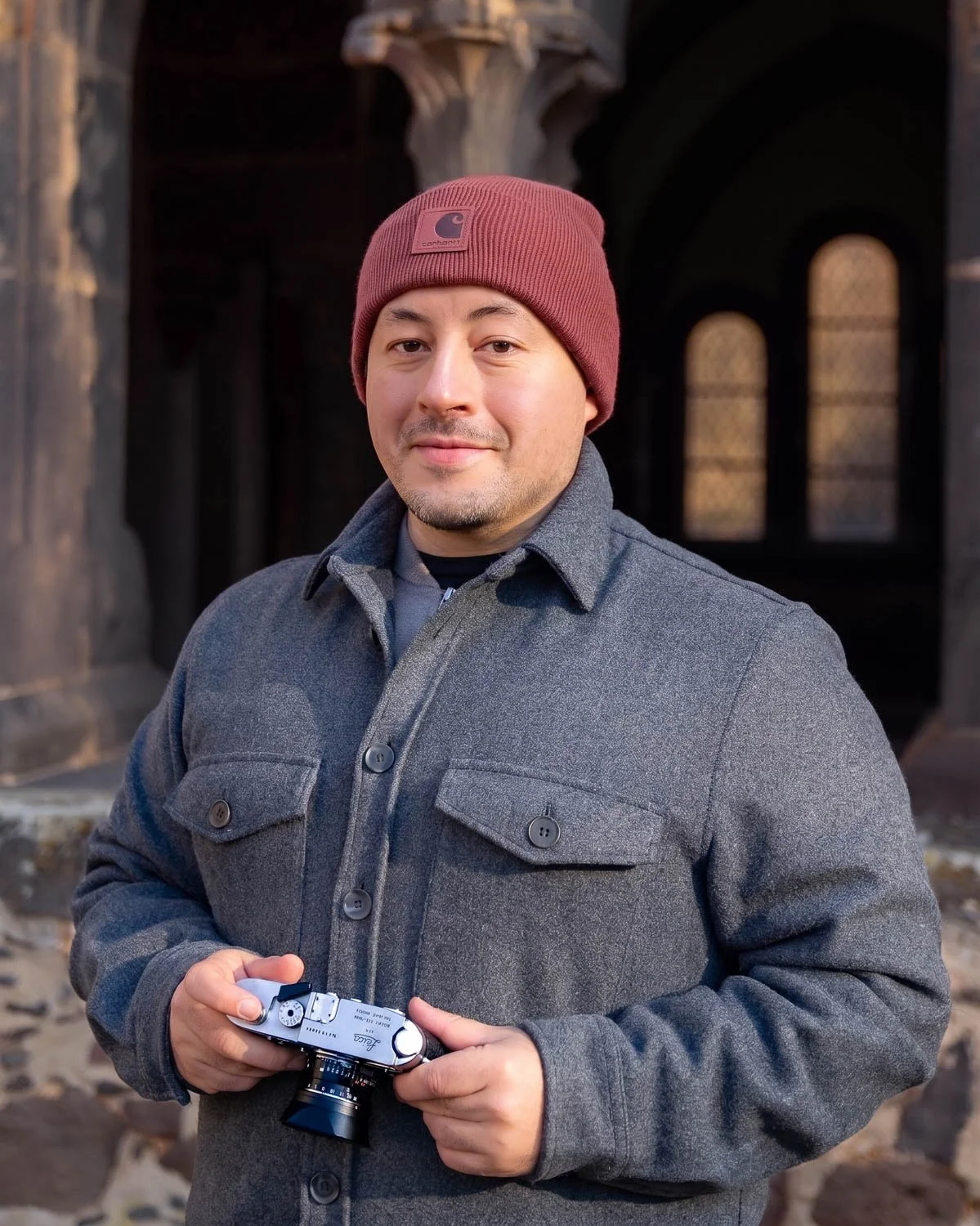 A man wearing a maroon beanie and gray coat holding a vintage camera, standing outdoors near a historic building with arched windows.