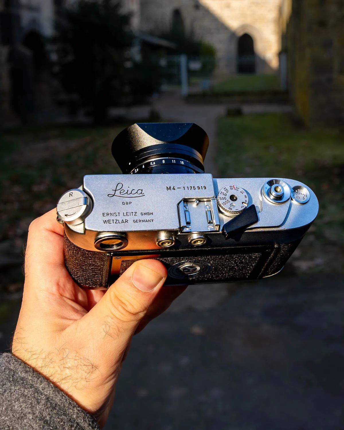 A person holding a vintage Leica camera outdoors with a blurred background of trees and a building.
