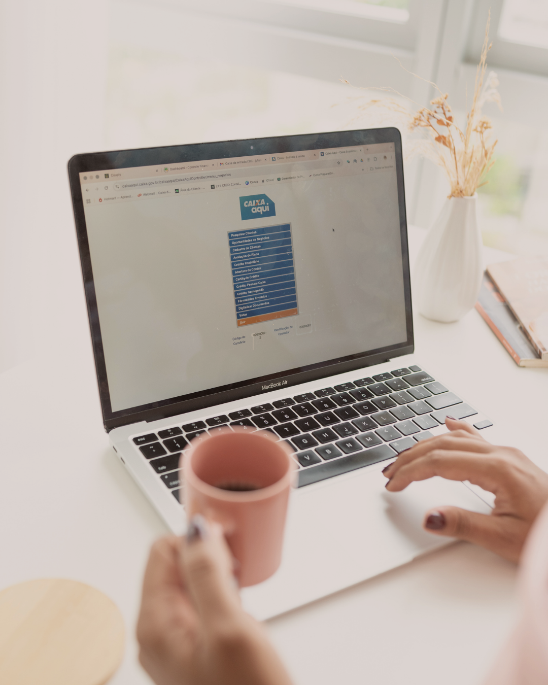 Pessoa usando um laptop MacBook Air com site do Banco Caixa Econômica Federal na tela, segurando uma xícara rosa na mão, em ambiente claro com vaso de flores e livros na mesa.