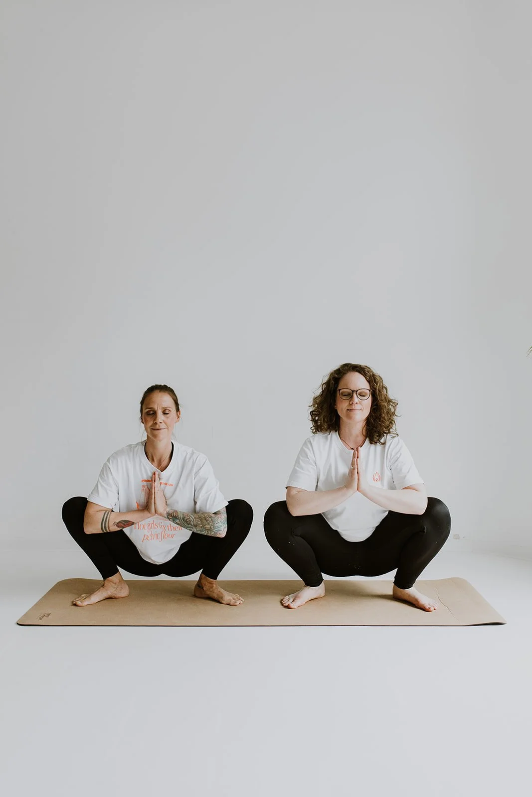 Zwei Frauen üben Yoga im Schneidersitz auf einer Matte, Hände vor der Brust in Gebetshaltung, in einem hellen Raum.
